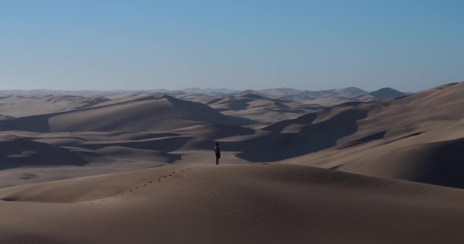 A distant view shows a man standing on the peak of a massive sand dune with endless rolling hills of sand stretching to the horizon under a clear blue sky