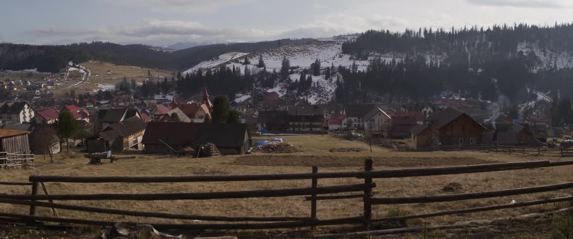Rural village with scattered houses wooden fence and hills partially covered with snow in the background