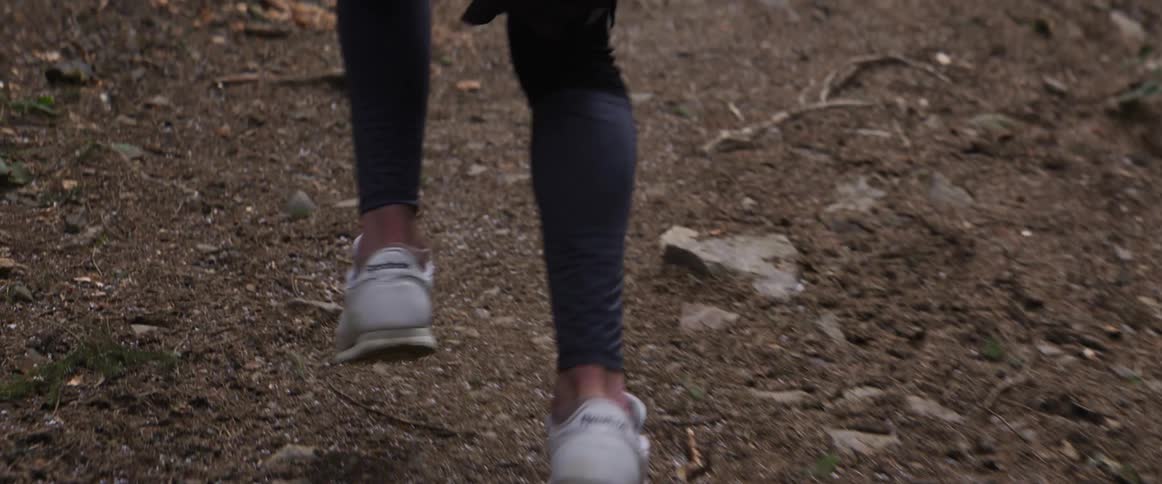 A woman wearing black leggings and white sneakers runs up a steep dirt trail filled with roots and stones in a forest