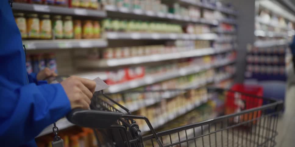 A person in a blue jacket pushes a shopping cart through a grocery store aisle while checking a small paper list the camera remains focused on their hands and the cart handle as they move past shelves stocked with various jars and food products