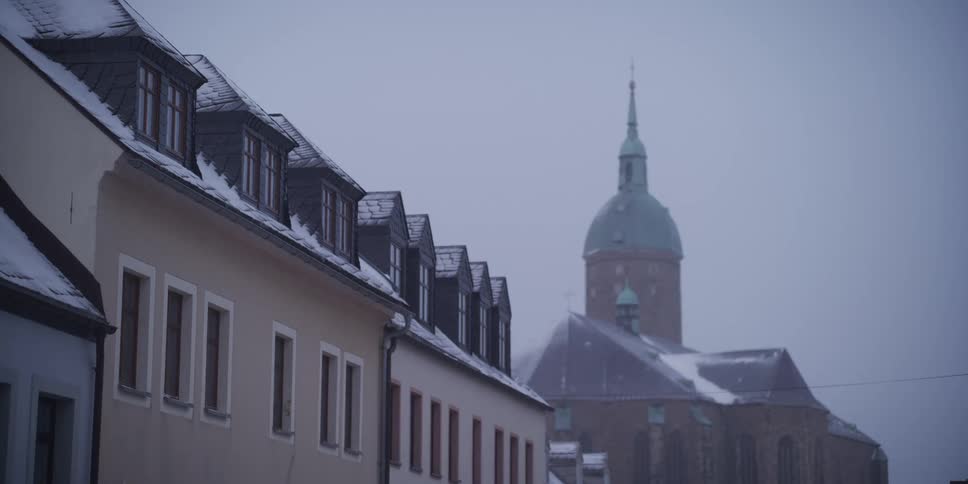 A view of snow covered roofs on residential buildings leads the eye toward the green dome and spires of saint annen church in annaberg buchholz under a thick and foggy winter sky