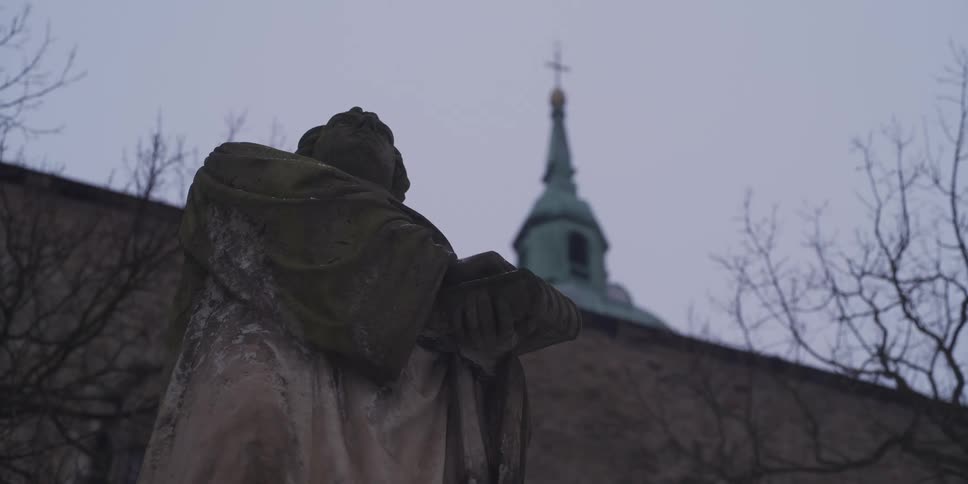 Statue with a religious theme stands near a building between leafless trees after dusk while a church towers in the background
