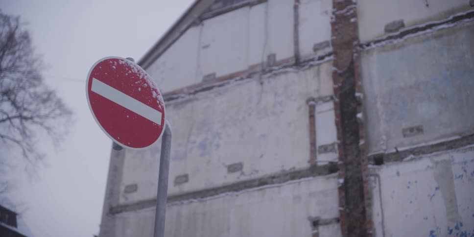 A red circular no entry road sign covered in light snow stands in front of a weathered and crumbling concrete building under a grey winter sky