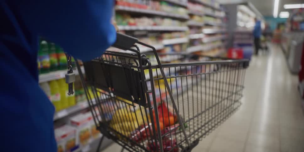A person in a blue jacket pushes a shopping cart containing bananas and other items through a grocery store aisle lined with various food products on shelves while another shopper walks in the blurred background