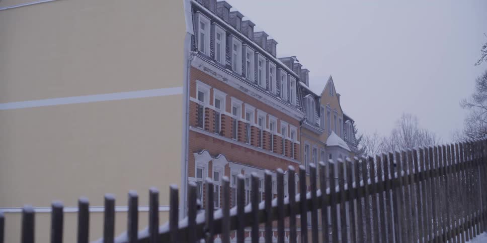 A multi story brick and yellow apartment building stands under a gray winter sky behind a dark metal fence covered in light snow while bare trees visible in the background