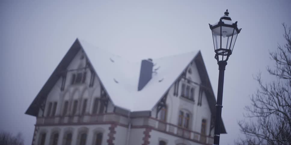 A street lamp stands in the foreground against a large european style villa with a snow covered gabled roof under a gloomy overcast sky during winter
