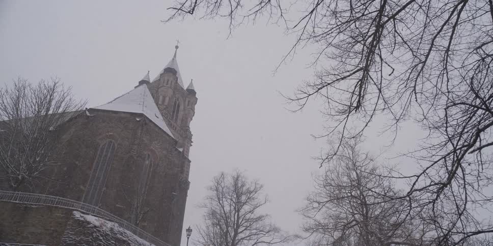 A tall stone church rises above snowdusted trees as light snow falls under a pale gray winter sky