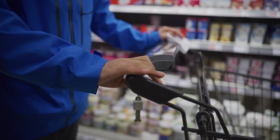 A person in a blue jacket pushes a shopping cart down a grocery store aisle lined with refrigerated products