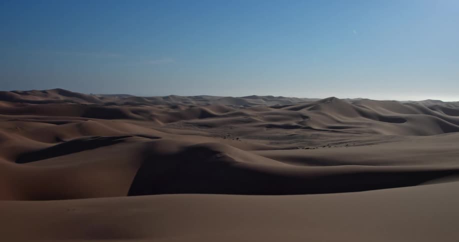 This aerial footage captures a vast expanse of rolling sand dunes stretching toward a clear bright horizon the shifting light creates deep shadows across the undulating desert landscape highlighting the natural textures of the terrain