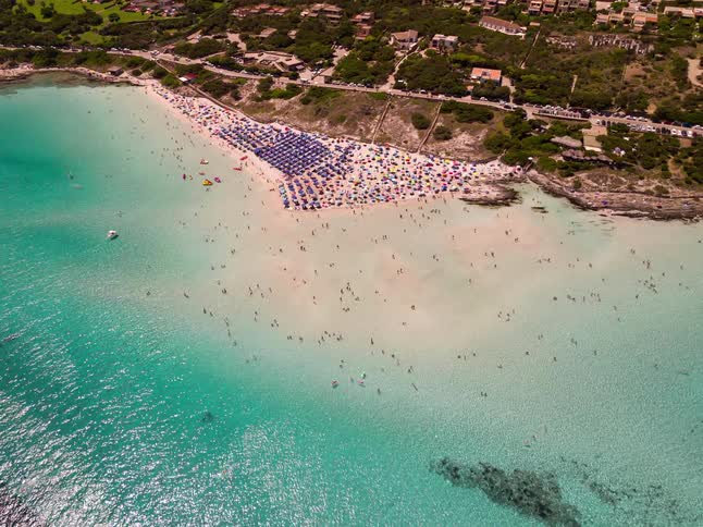 Ariel view of a crowded beach with vibrant turquoise water bordering a coastal town and hilly landscape