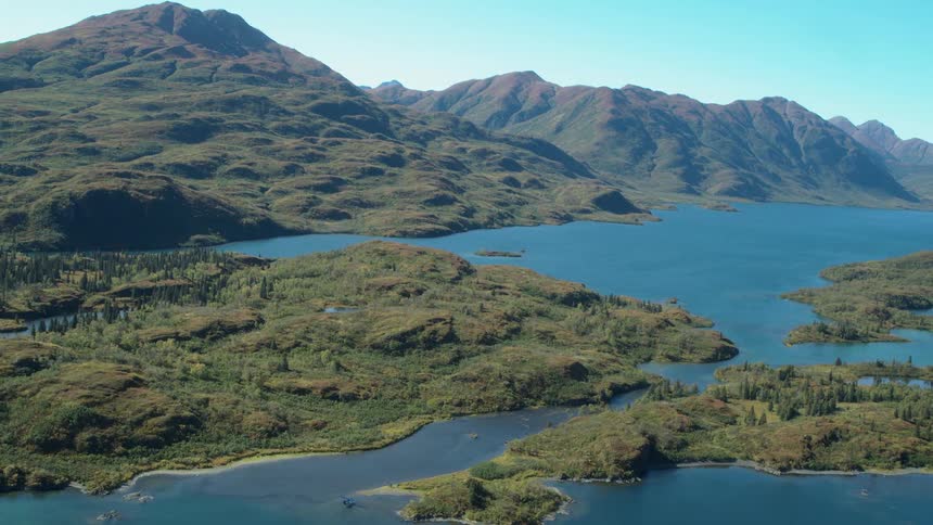 An aerial shot shows a vast blue lake winding through green hills and lush vegetation under a clear sky with large mountain ranges