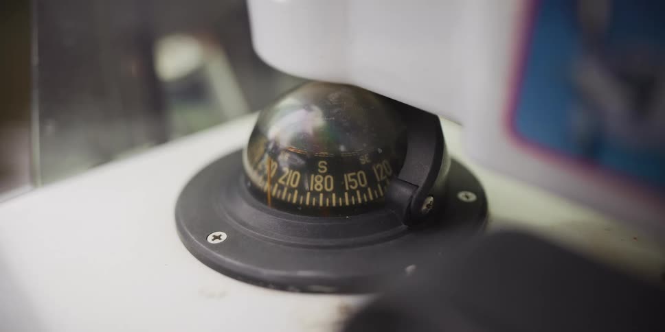 A nautical compass sits mounted on a vessel its dome slightly weathered and reflecting the surrounding light the yellow markings on the dark dial indicate headings as the ship moves through the water