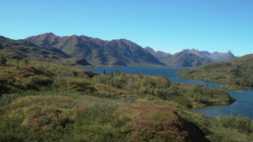 The video features an aerial tracking shot over a lush green wilderness with a flowing river and distant mountains as the camera moves forward it reveals a vast deep blue lake nestled within the rolling hills under a clear sky