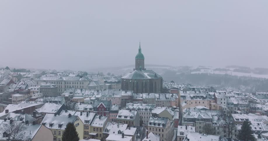 The video shows a wide aerial view of a european town blanketed in thick snow under a heavy overcast skya large church with a distinct green copper dome and a tall spire stands as the central landmark surrounded by rows of colorful buildi