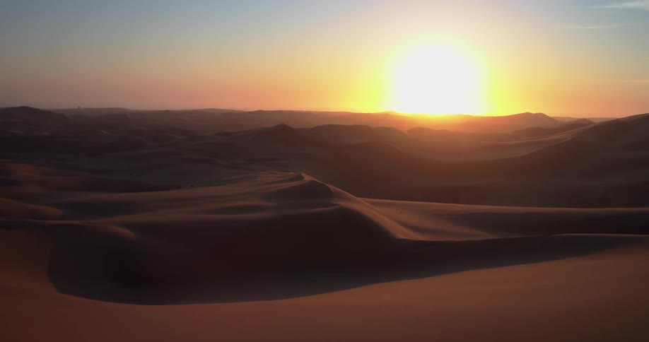 This aerial footage captures a vast desert landscape at sunset with golden light illuminating the peaks of rolling sand dunes the camera slowly pans across the undulating terrain emphasizing the deep shadows and the serene desolate beauty of the envir