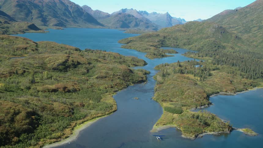An aerial view shows the vast blue water of a lake surrounded by green hilly terrain and rugged mountains as a small floatplane sits near a narrow channel under a clear sunny sky