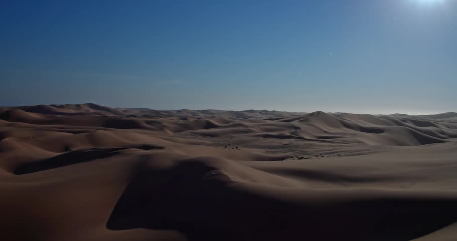A wide aerial view reveals the vast and undulating sand dunes of the namib desert under a clear blue sky as the bright sun creates deep shadows across the desert landscape