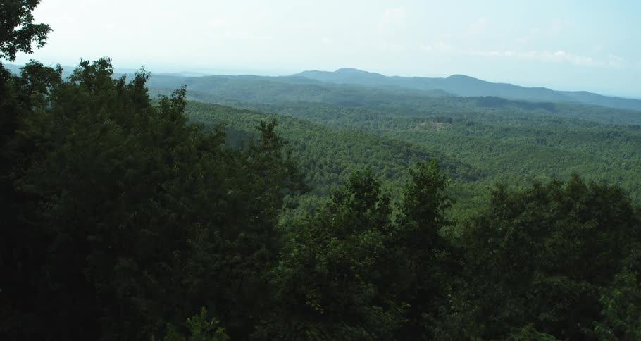 A wide view from a high elevation reveals the rolling green hills and dense forests stretching toward a distant horizon under a bright and clear summer sky