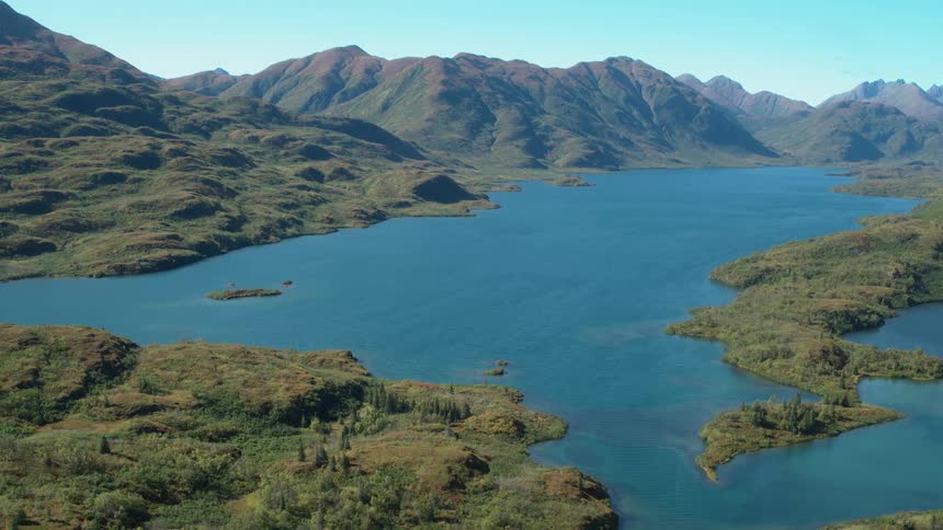 An aerial view shows the vast blue water of a lake surrounded by green hilly terrain and rugged mountains on kodiak island in alaska under a clear sunny sky