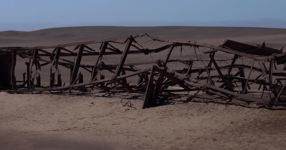 A view from the side shows a long rusted and skeletal metal structure lying collapsed on the sand of the skeleton coast in the namib desert under a clear blue sky