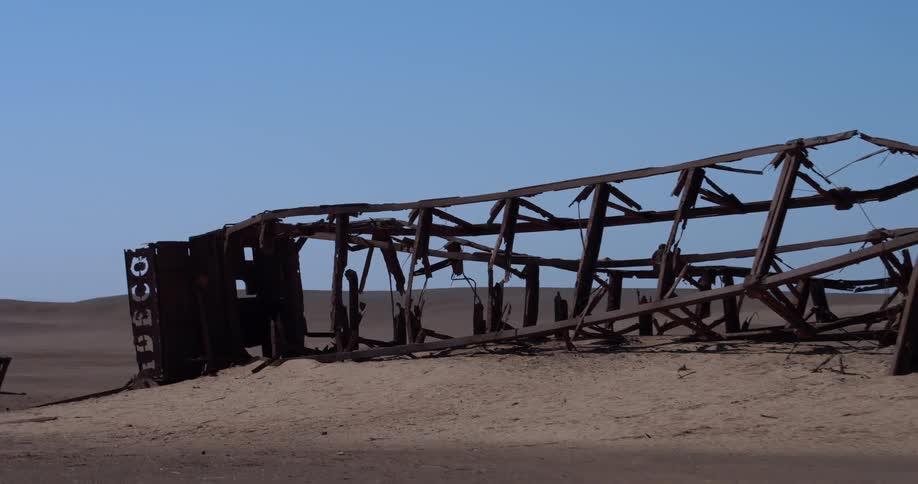 A rusted shipwreck lies partially buried in sand in a barren desert landscape under a clear sky