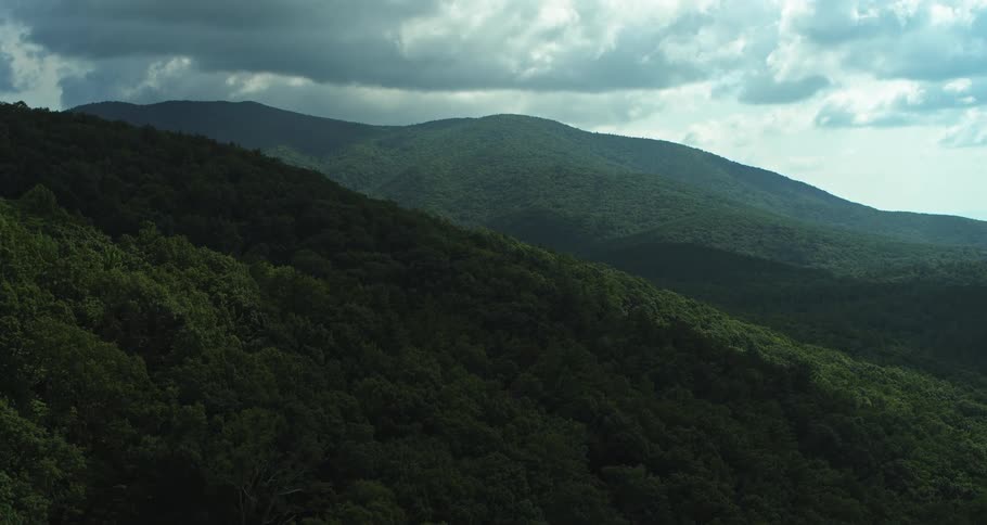 The video features a slow sweeping aerial shot of lush green mountain ridges under a dramatic cloudy skylight and shadow play across the dense forest canopy creating a moody and expansive atmosphere as the camera glides over the peaks