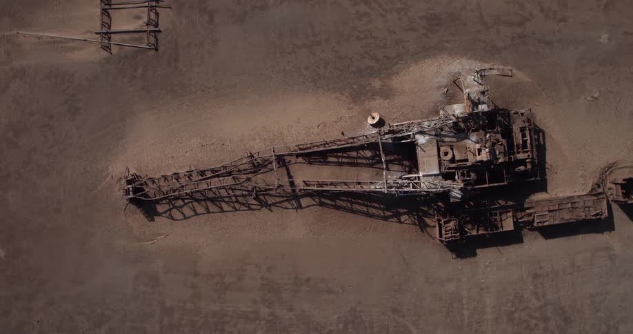 An overhead drone shot captures a massive rusted industrial excavator abandoned in the middle of a barren desert where the dry ground shows tracks and scattered metal remnants from the decaying machine