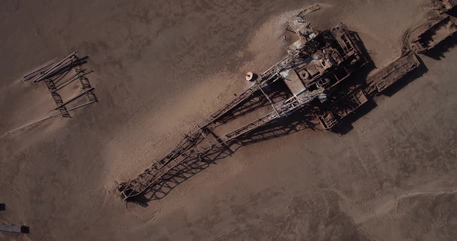 A top down aerial shot captures a massive rusted excavator lying abandoned in a vast sandy desert
