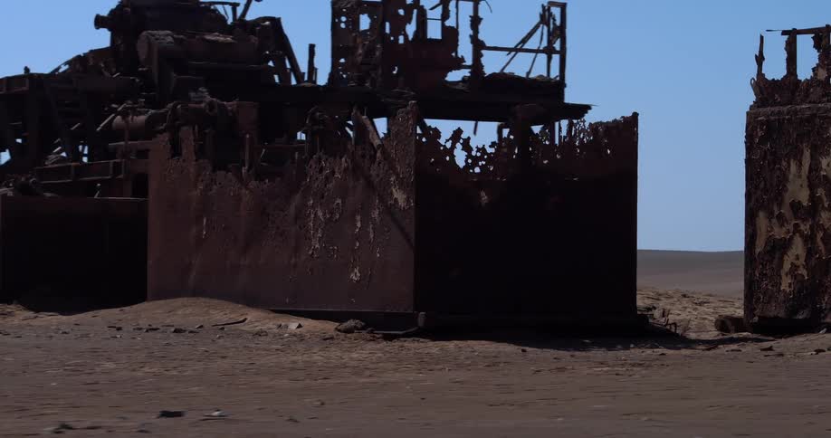 A massive rusted excavator bucket sits halfburied in sand beneath the skeletal remains of an abandoned industrial structure