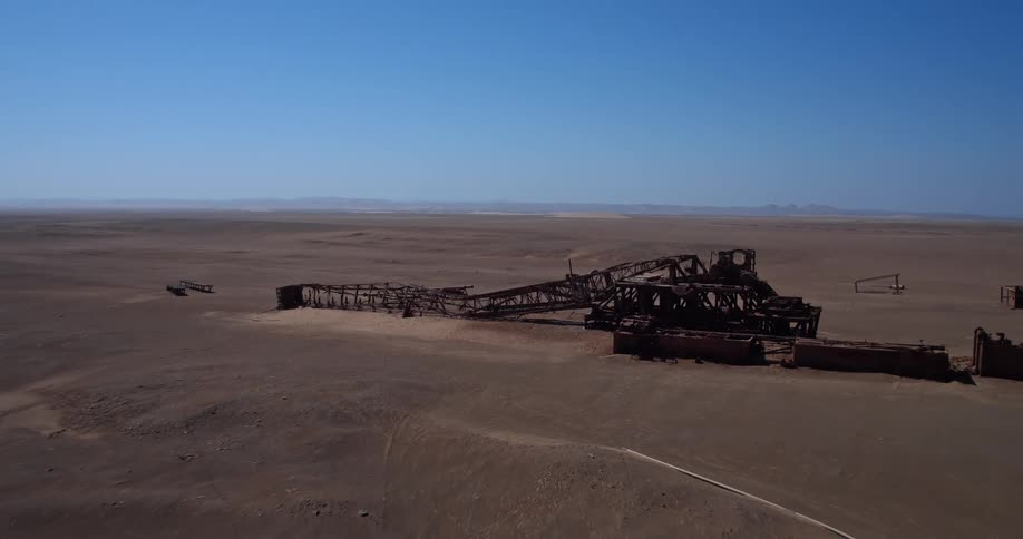 An aerial shot shows a large rusted industrial structure abandoned in a vast desert landscape under a clear blue sky