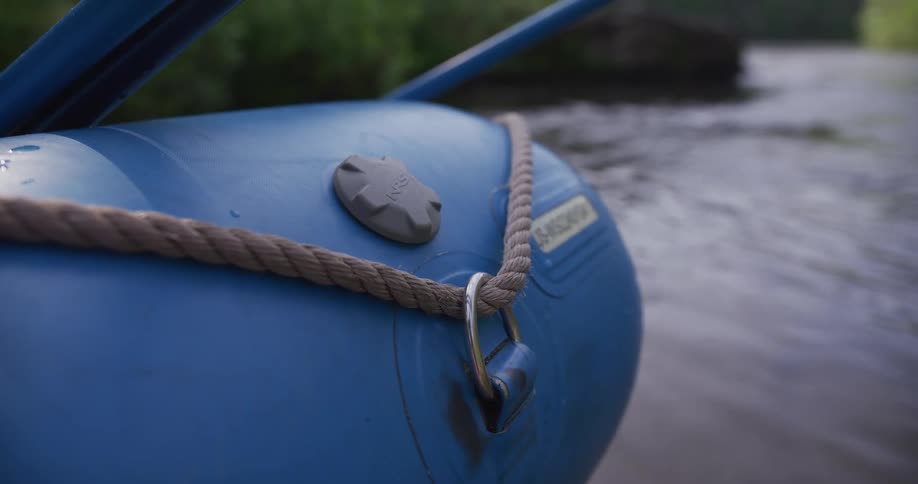 A close view shows a thick rope secured to a metal ring on a blue inflatable raft floating on a river with dark water and green trees in the background