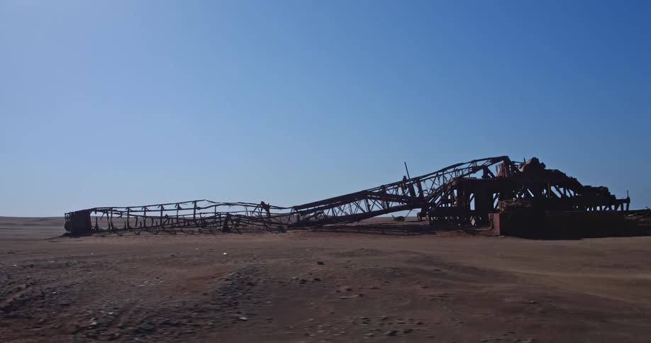 A rusted and collapsed industrial metal structure sits abandoned in the middle of the namib desert at the skeleton coast in namibia under a clear blue sky