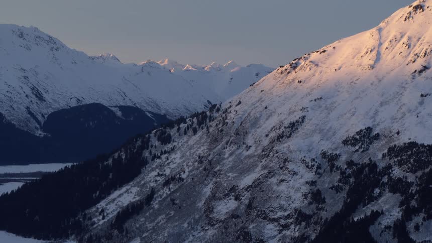 Golden sunlight hits the snow covered slopes of the chugach mountains in alaska above a dark forest and a frozen valley during a cold winter sunset