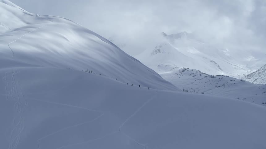 A distant view shows a group of hikers walking in a line across a steep snow covered mountain ridge in the alps under a cloudy and bright winter sky
