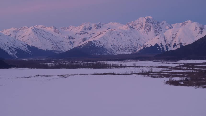 Soft pink and purple light from a winter sunset glows over the snow covered peaks of the chugach mountains above a vast frozen valley in alaska