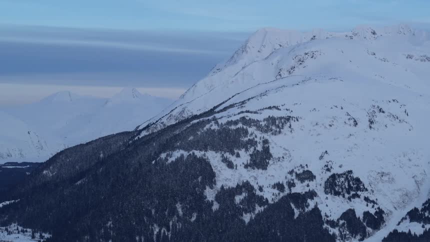A winter landscape of a mountain with snow covering most of its formation with grasses and trees on parts of its slope at sunrise
