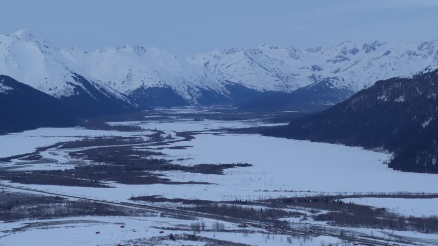 A wide aerial view of the frozen matanuska river valley in alaska with snow covered plains and dark forests leading toward a distant range of jagged white mountains under a pale sky