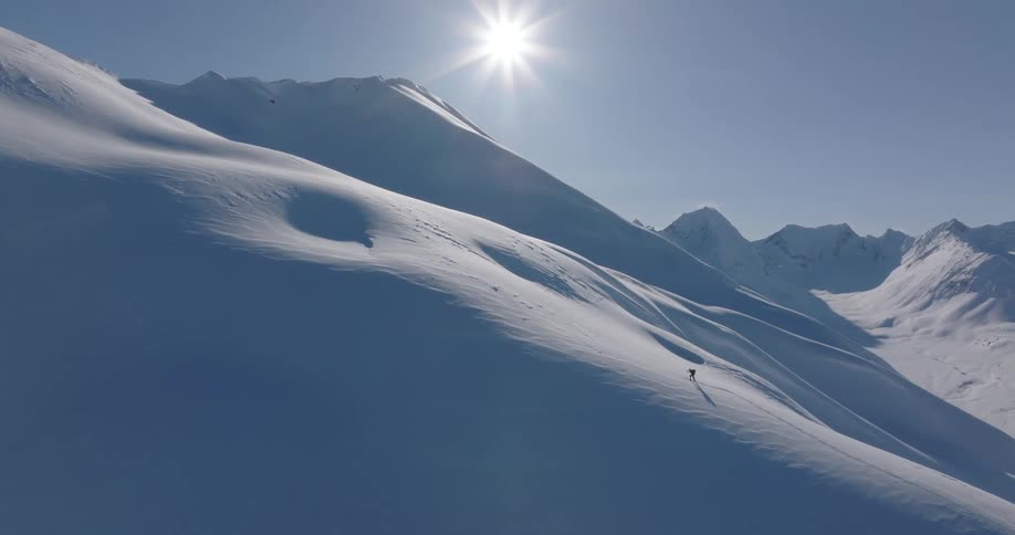 A person treks across a steep snow covered mountain ridge in bright sunlight