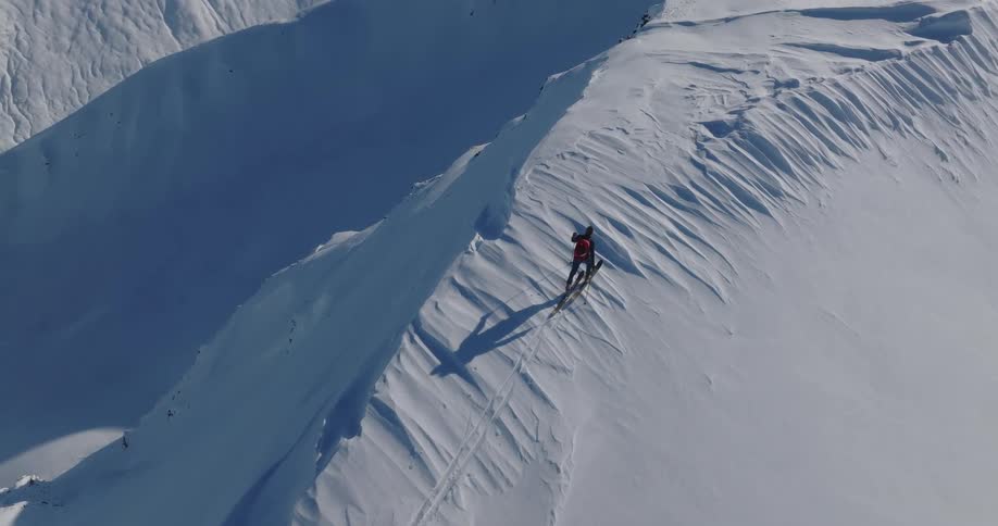 A lone skier in a red jacket glides along a sharp snowy ridge casting a long shadow across the untouched white slope