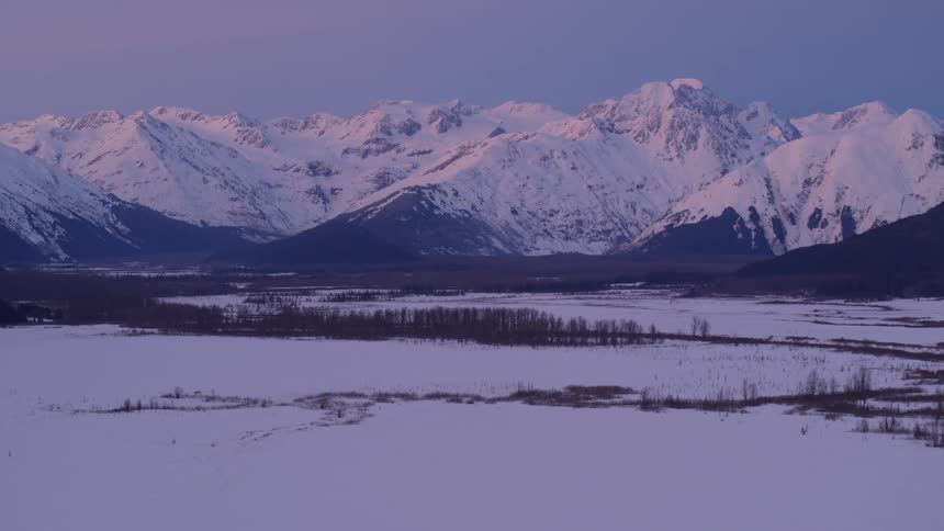 Purple and pink light from a winter sunset glows over the snow covered peaks of the chugach mountains above a wide frozen valley and dark forests in alaska