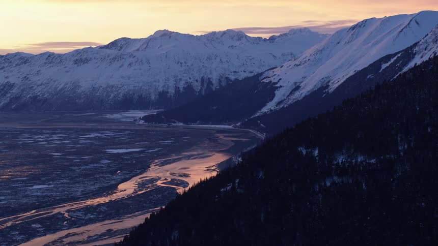 A winding river reflects the golden glow of sunset as it curves through a vast valley beneath towering snowcovered mountains