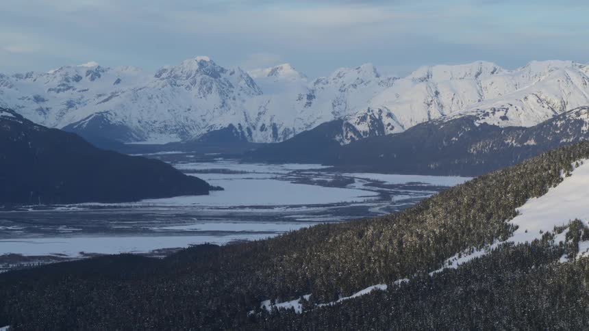 A wide aerial view shows the snowy peaks of the chugach mountains overlooking a frozen river valley and dark evergreen forests in alaska under a soft winter sky
