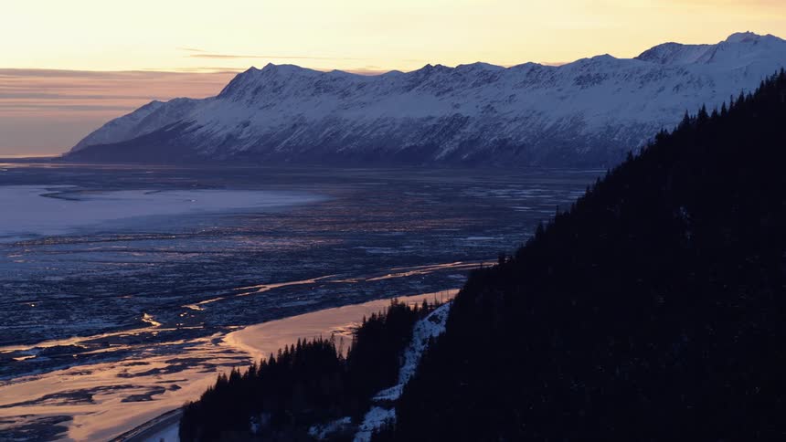 A glowing river winds through a vast valley beneath a range of snowcovered mountains as the sky fades into warm sunset tones