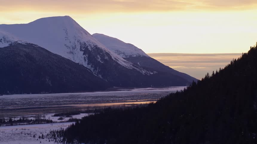 An aerial view shows a vast mountain range in alaska covered in deep white snow under a clear blue sky with bright sunlight creating shadows on the steep slopes and ridges