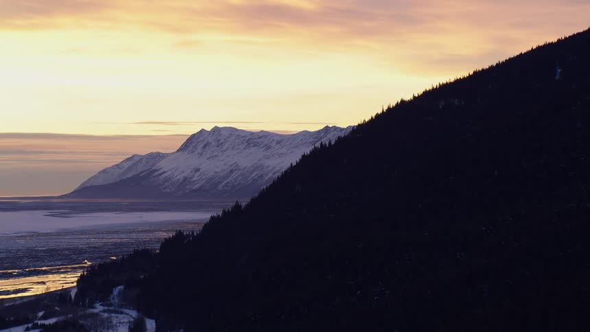 A mountain slope is covered with trees while snow covers the land below