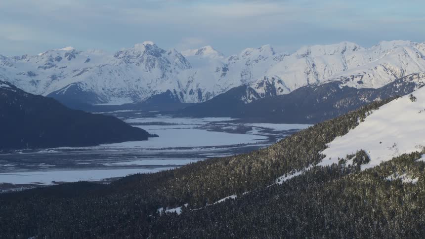 A wide aerial view shows the snowy peaks of the chugach mountains overlooking a frozen river valley and dark evergreen forests in alaska under a soft winter sky