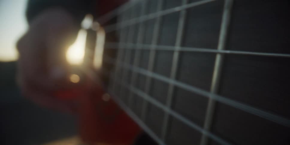 A close view shows the hands of a man playing a wooden acoustic guitar outdoors with bright sunlight shining through the strings and creating a warm flare