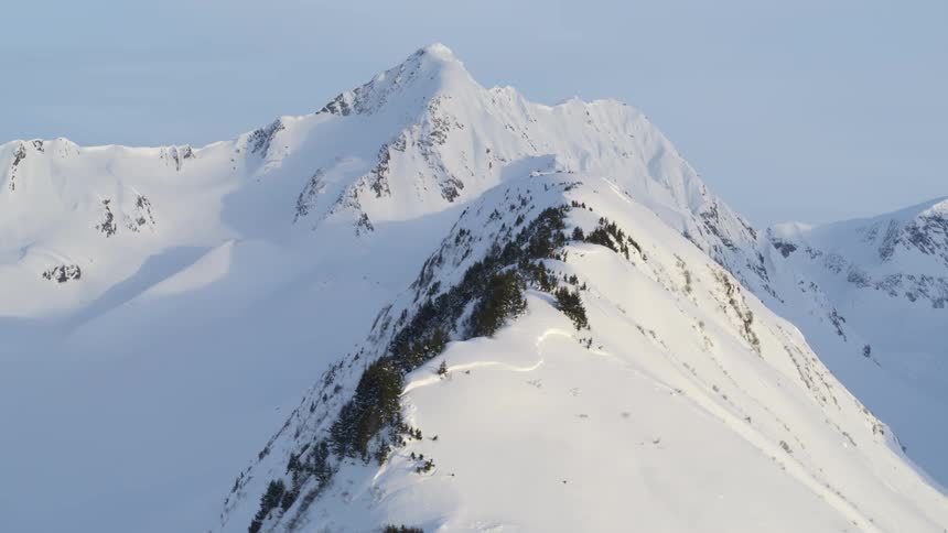 The video shows a sharp snowcovered mountain peak standing prominently against a clear sky a secondary ridge in the foreground is dotted with dark evergreen trees creating a stark contrast against the white slopes