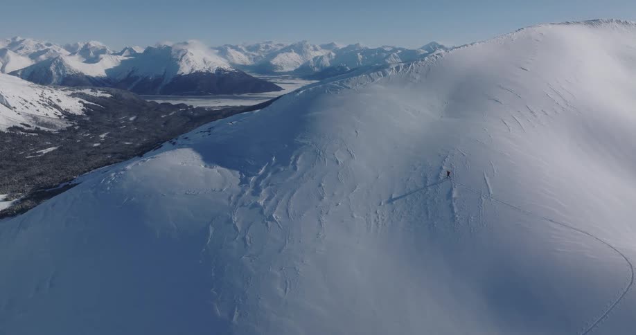 An aerial shot shows a person standing on a snow covered mountain ridge within a vast mountain range