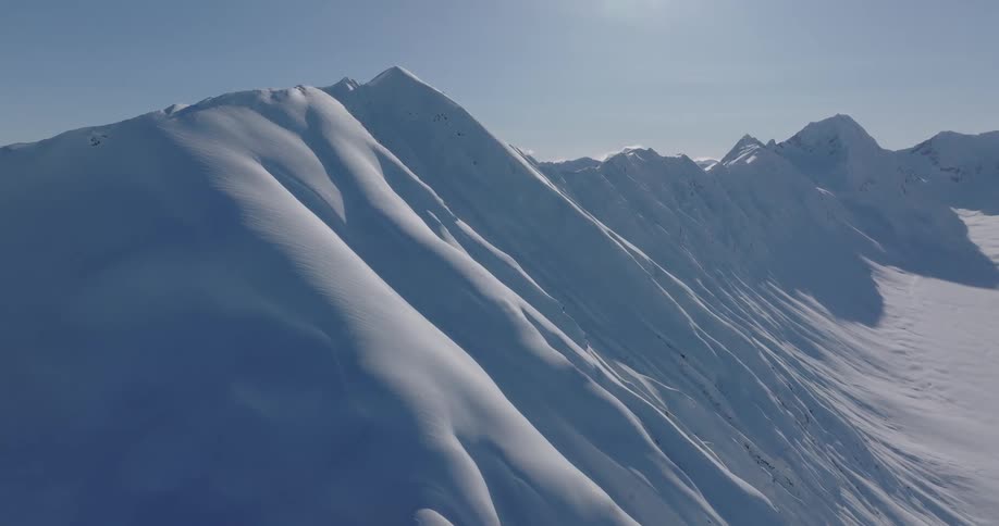 An aerial view shows a vast mountain range in alaska covered in deep white snow under a clear blue sky with bright sunlight creating shadows on the steep slopes and ridges